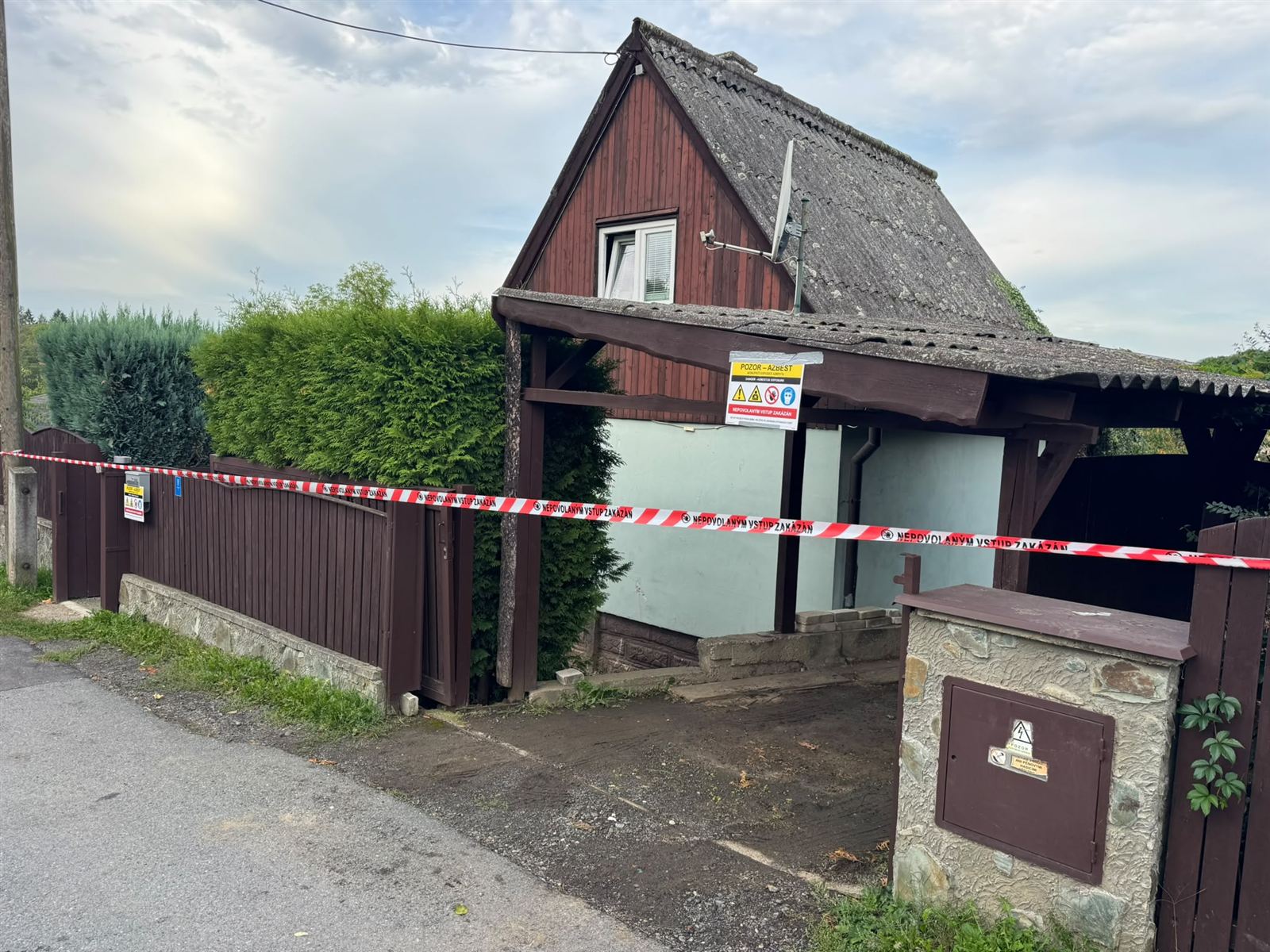 Dům s vlnitou eternitovou střechou před zahájením demontáže, na plotě výstražná páska s upozorněním na azbest
